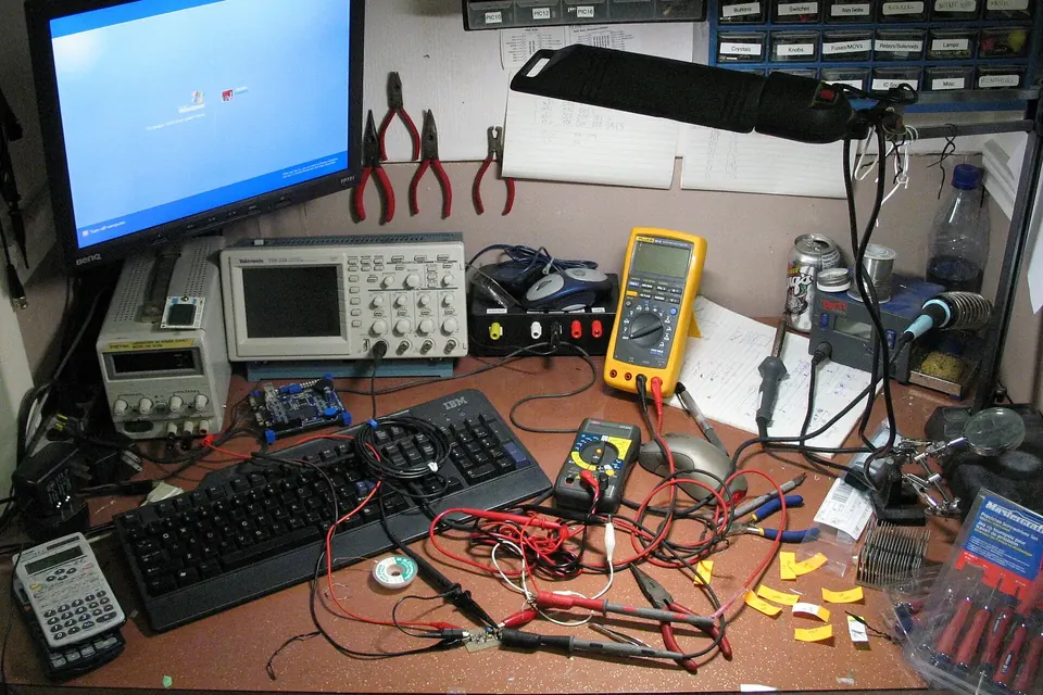 A cluttered electronics workbench with two digital multimeters, an oscilloscope, a bench power supply, test leads and a computer monitor